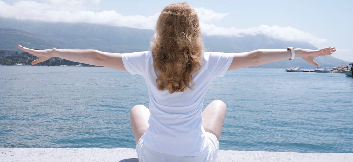 Woman sitting with open arms by the water, embracing nature and serenity.