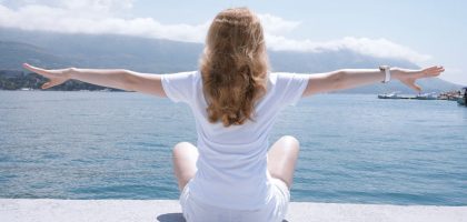 Woman sitting with open arms by the water, embracing nature and serenity.