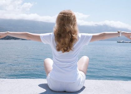Woman sitting with open arms by the water, embracing nature and serenity.